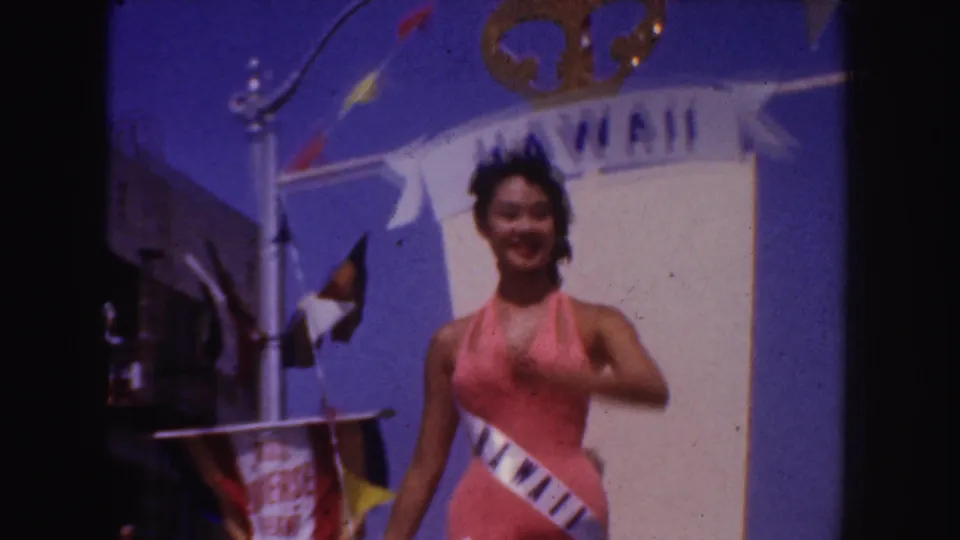 A woman in a pink swimsuit and white sash stands smiling on a Hawaii-themed parade float, with street details at the edge of frame.