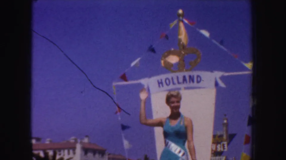 In 1952 California home-movie footage, a woman in a blue swimsuit and sash waves from a float labeled Holland.