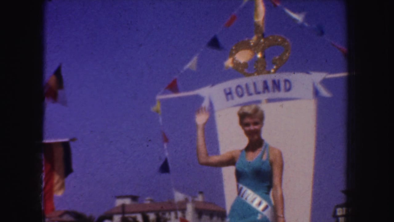 Blonde woman in blue swimsuit waves beneath a HOLLAND banner during a parade.