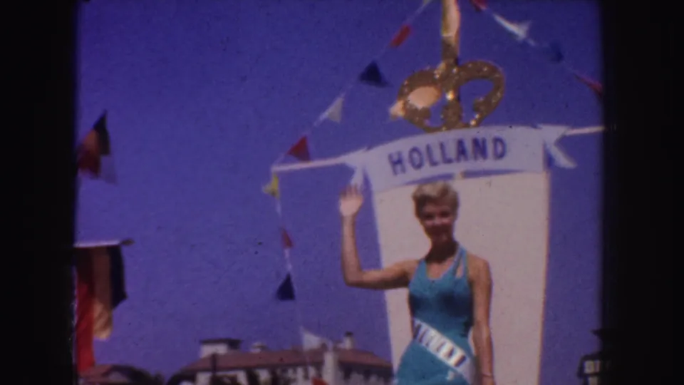 In 1952 California parade footage, a blonde woman in a blue swimsuit and sash waves beneath a HOLLAND banner with a crown decoration.