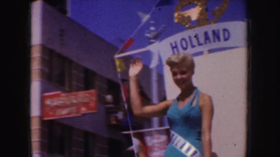 The waving woman stands beside the HOLLAND parade display, with small flags, a crown ornament, and street buildings visible behind her.