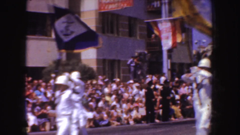 Uniformed men march past a large seated crowd on a California street, with parade signage and building facades in the background.