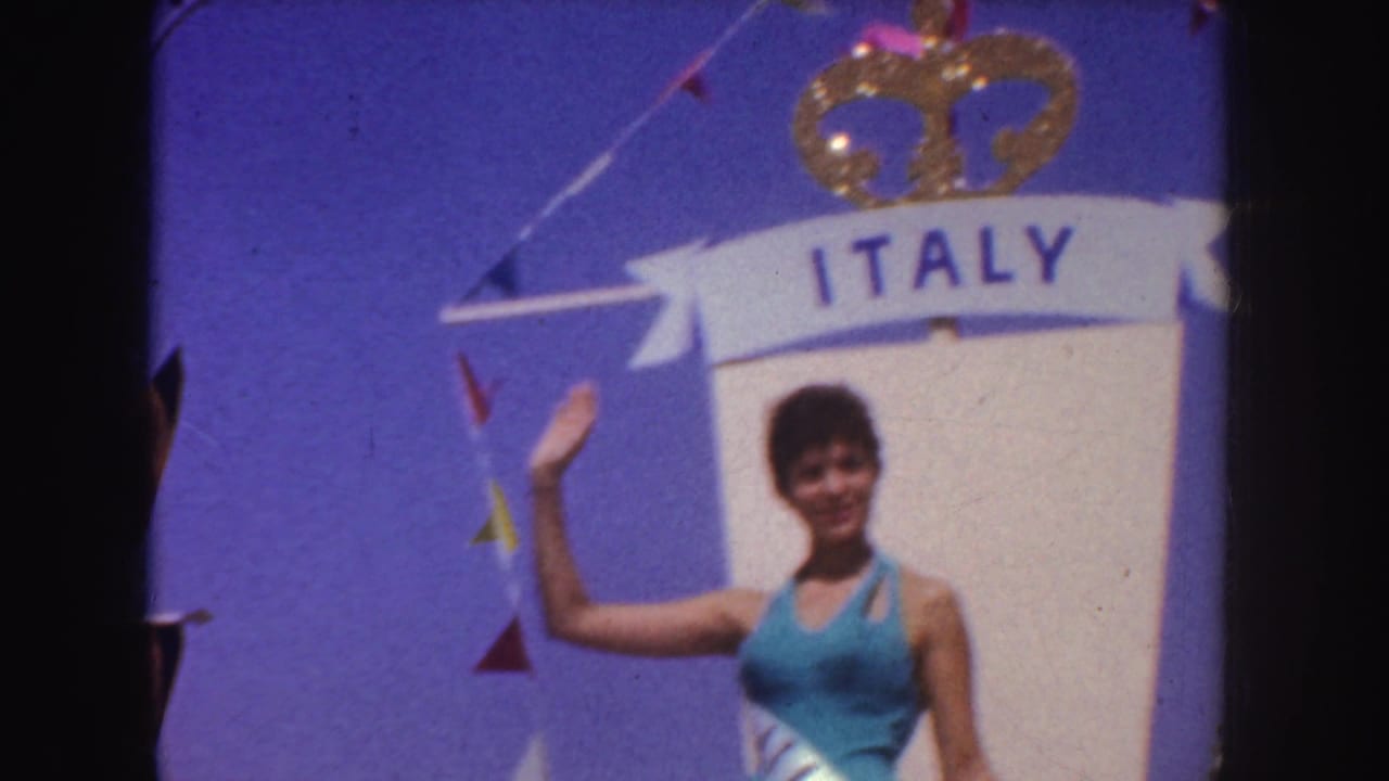 Woman in a blue dress waves beside a large ITALY float sign with a gold crown.