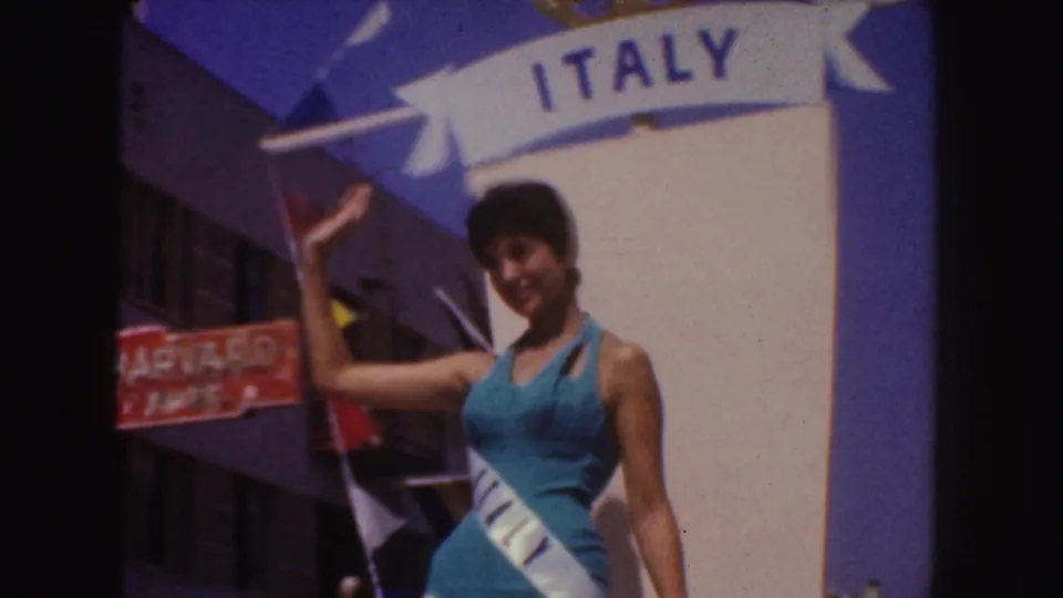 Woman in blue waves from an Italy-themed float during a 1952 California parade, with storefront signage and buildings behind her.