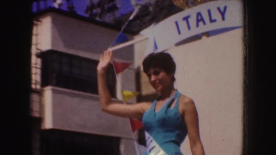 Closer 8mm view of the woman in a blue dress and sash waving under the ITALY sign on the parade float.