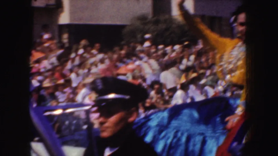 1952 California parade frame with a police officer in the foreground, a costumed participant, and spectators seated behind them.