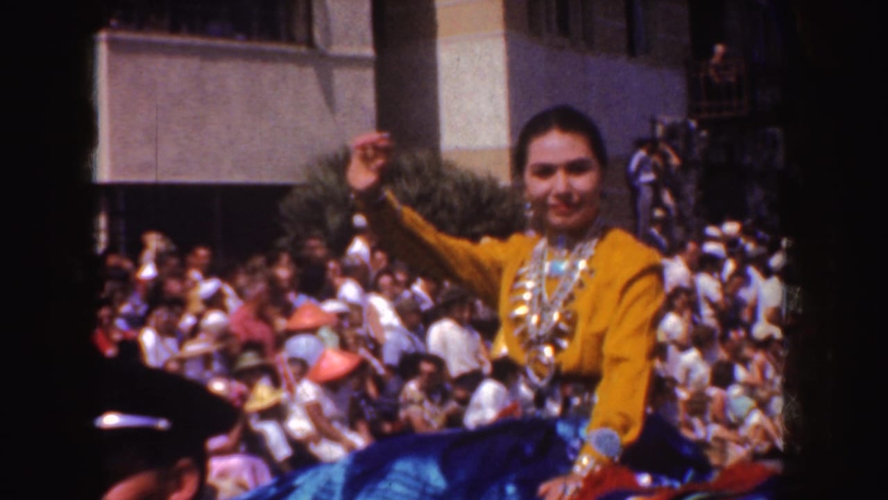 Woman in yellow waves from a parade float beside a crowded sidewalk.