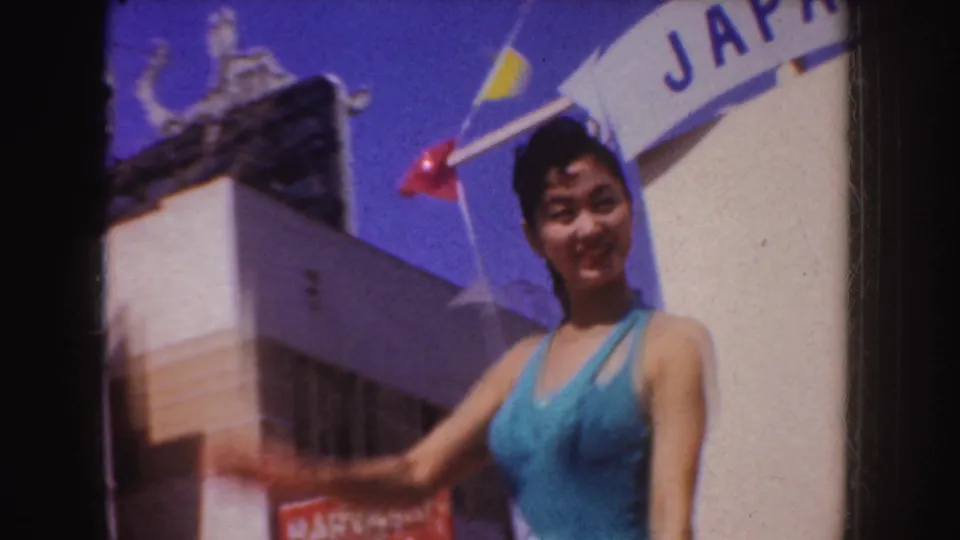 A woman in a blue swimsuit-style outfit waves from a parade float near a visible Japan banner in the supplied 1952 California footage.