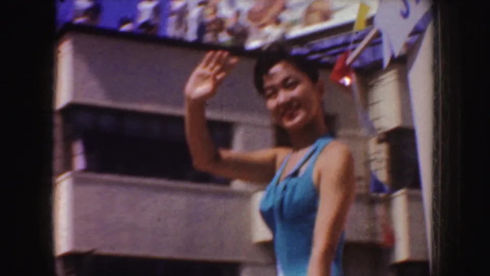 A smiling woman in blue waves from a float, with a building facade and partial parade decorations behind her in the 1952 California clip.