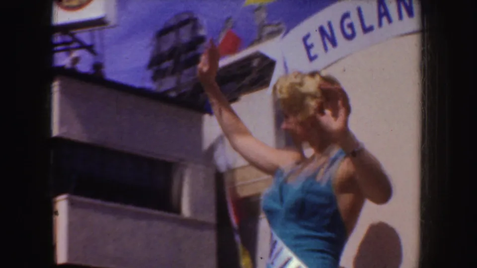A blonde woman in a blue outfit waves from a float below a curved England banner during the supplied 1952 California parade footage.