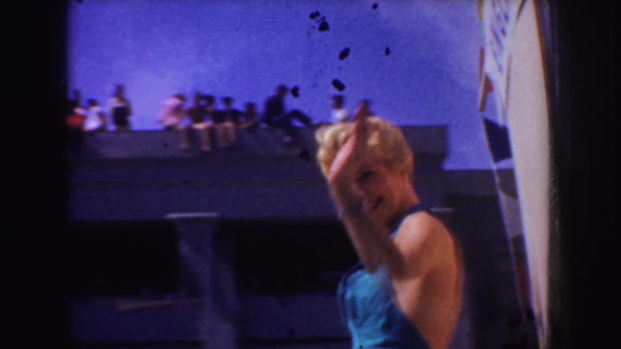 In 1952 California home-movie footage, a blonde woman in teal parade attire smiles and waves near a ship-like structure under blue sky.