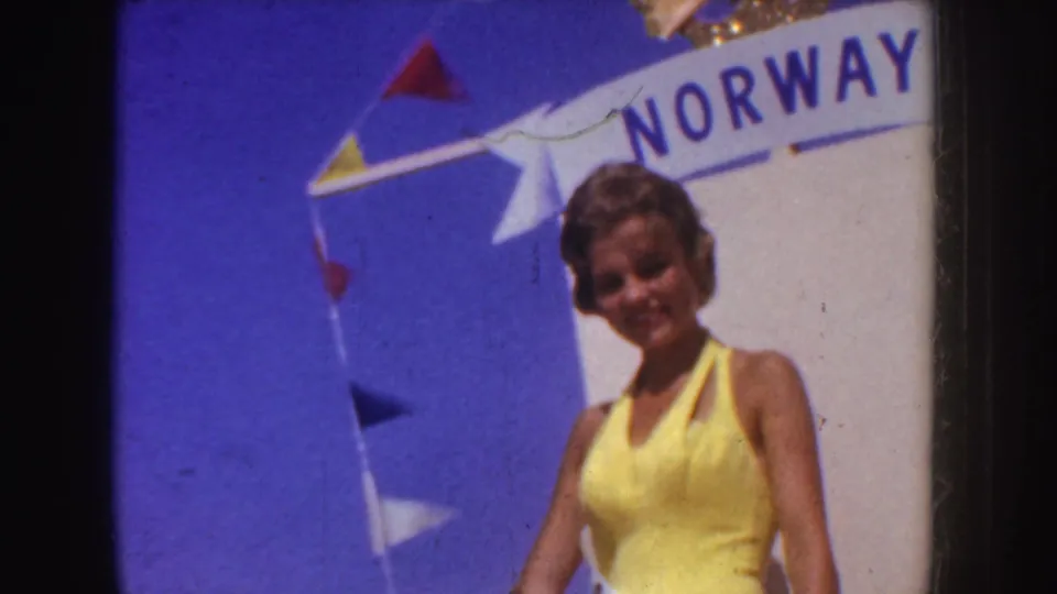 A woman in a yellow halter dress poses near a visible Norway sign and colorful pennant flags in 1952 California parade footage.