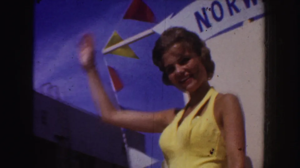 The woman in yellow waves toward the camera beside the Norway sign, with triangular pennants visible against the blue sky.