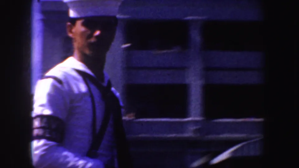 A sailor in a white uniform and cap passes through the frame near a large ship-like structure in the 1952 California parade footage.