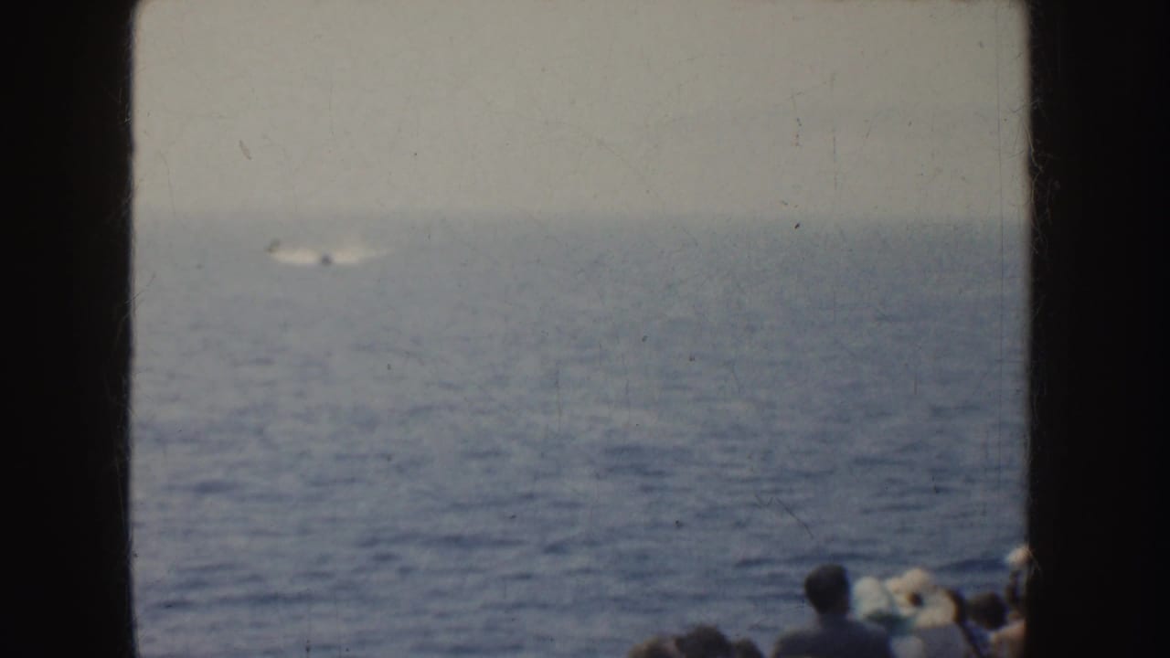 1952 California 8mm frame showing a small motorboat far offshore, leaving a pale wake as people watch from the foreground.