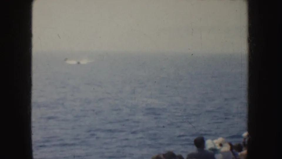 1952 California 8mm frame showing a small motorboat far offshore, leaving a pale wake as people watch from the foreground.