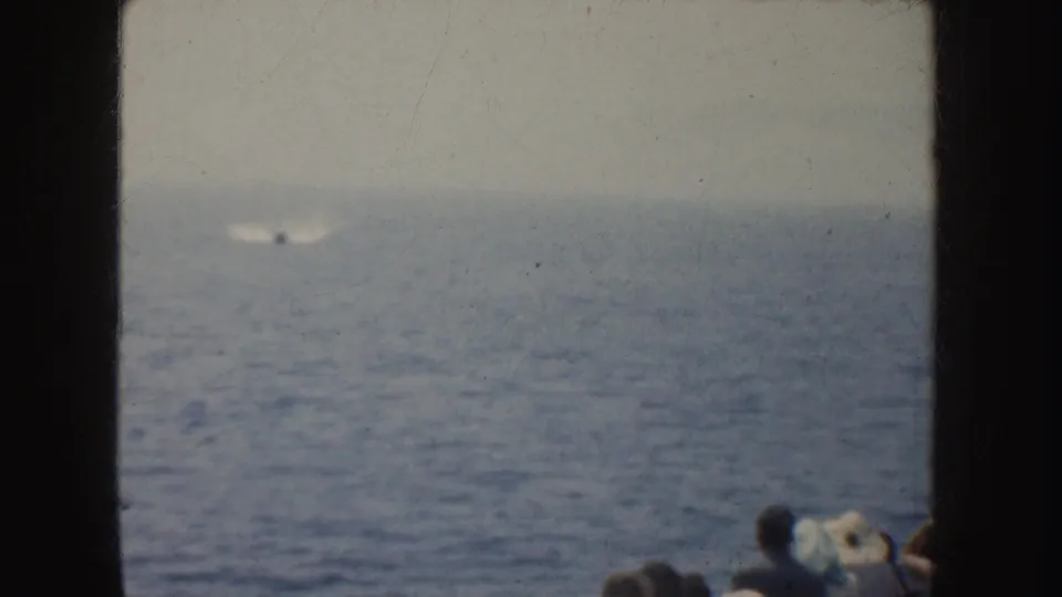 1952 California home-movie frame with a faint motorboat and wake offshore, seen past foreground spectators and dark film borders.