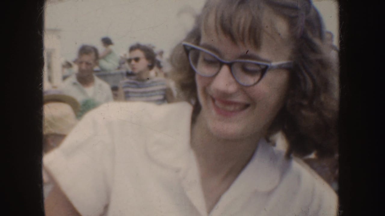 A smiling woman with cat-eye style glasses sits among outdoor spectators in 1952 California home-movie footage.