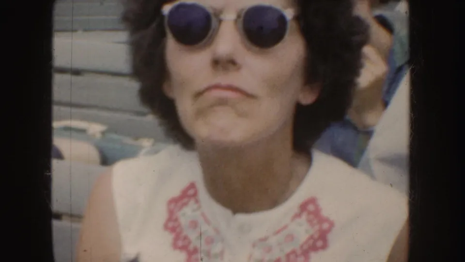 In 1952 California footage, a woman with dark curly hair and round sunglasses looks toward the camera in a white embroidered blouse.