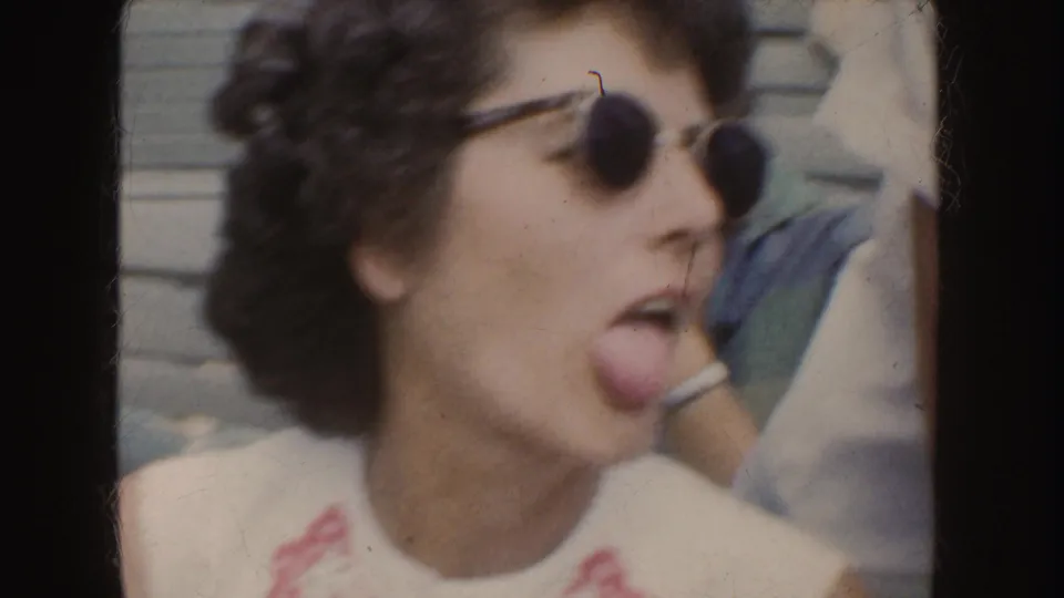 In a playful 1952 California home-movie frame, a woman in round sunglasses sticks out her tongue near other seated people.