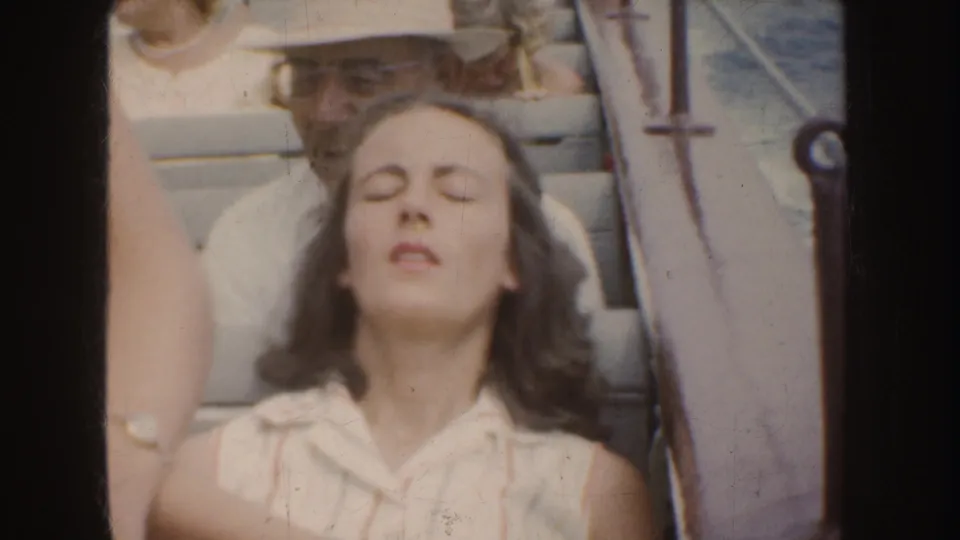 In 1952 California home-movie footage, a long-haired woman in a sleeveless light blouse sits with eyes closed among people outdoors.
