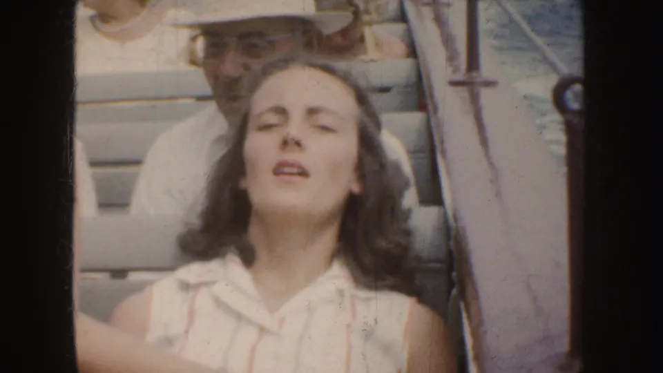 In 1952 California home-movie footage, a young woman on a boat tilts her face upward with eyes closed as a man sits behind her.