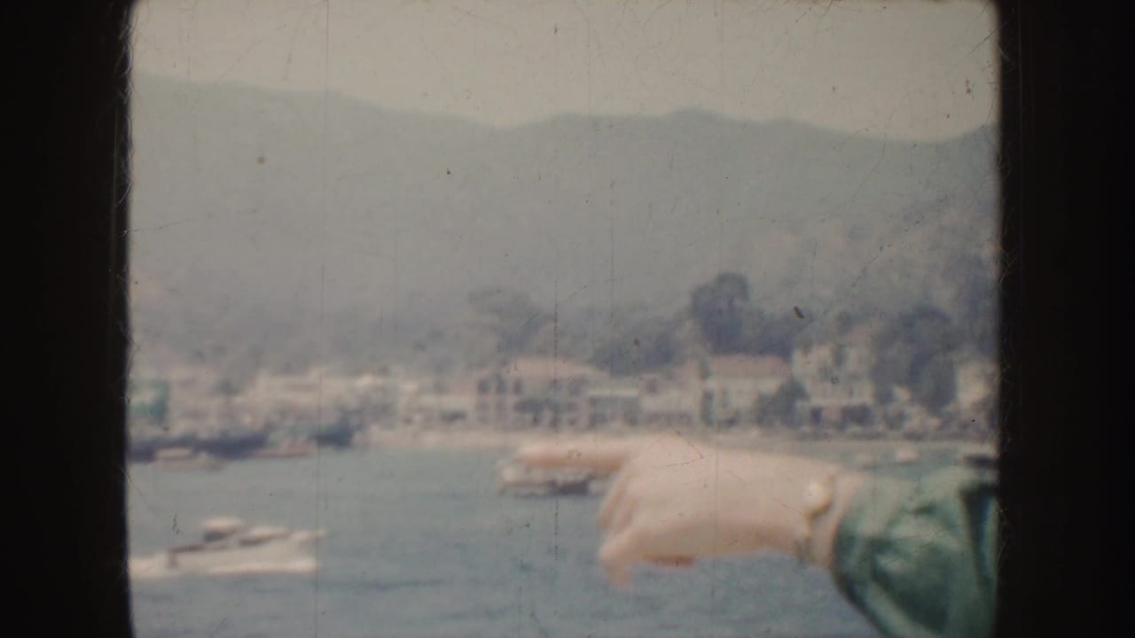 1952 California home-movie frame with a hand pointing toward boats in a coastal harbor below pale hillside buildings.