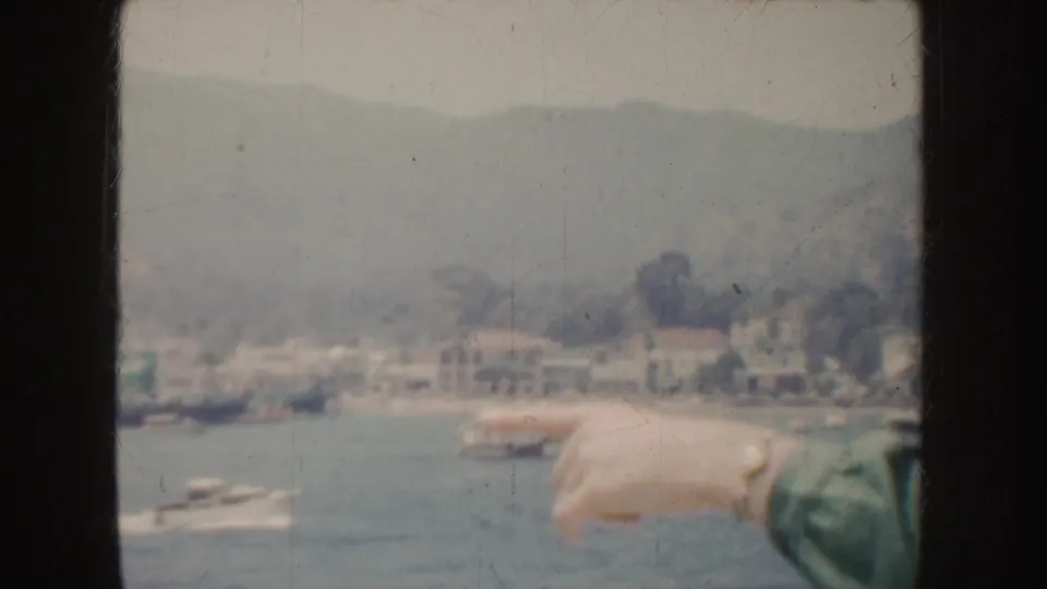 1952 California home-movie frame with a hand pointing toward boats in a coastal harbor below pale hillside buildings.