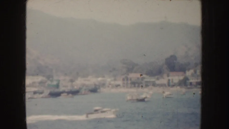 Soft 8mm view of a California coastal harbor, with small boats, wakes, waterfront buildings, and hills in the distance.