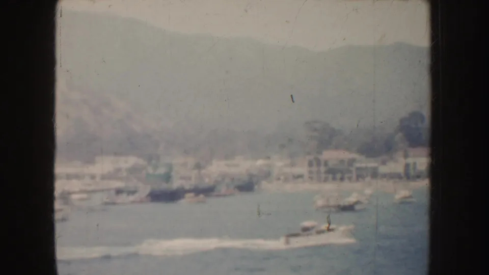 1952 California harbor view with boats and a bright wake below soft-focus waterfront buildings and rolling hills.