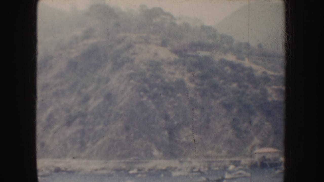Hazy wooded hillside above calm waterfront with small boats and shoreline structures.