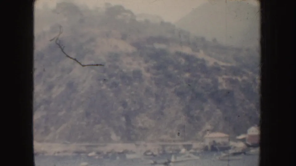 Distant 1952 California waterfront view with a wooded mountain slope, small boats, and low buildings along the shore.