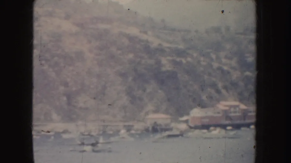 1952 California 8mm frame of a calm waterfront, clustered small boats, shoreline structures, and a softly faded hillside beyond.