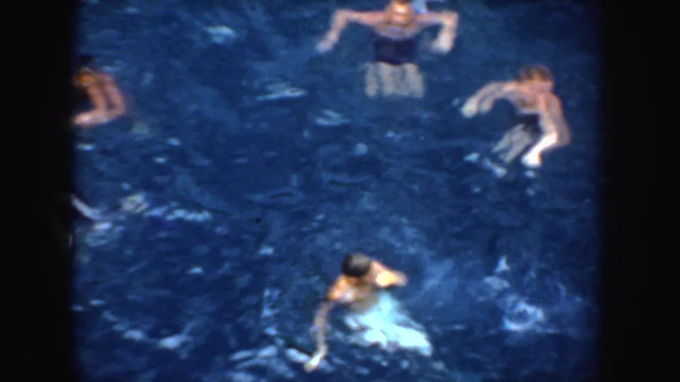 Several swimmers are visible in blue water, including one near a pale object, in a soft 8mm home-movie frame.