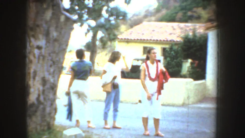 Three adult women in light summer clothing stand outside in California, near a tree, low wall, and tiled-roof building.