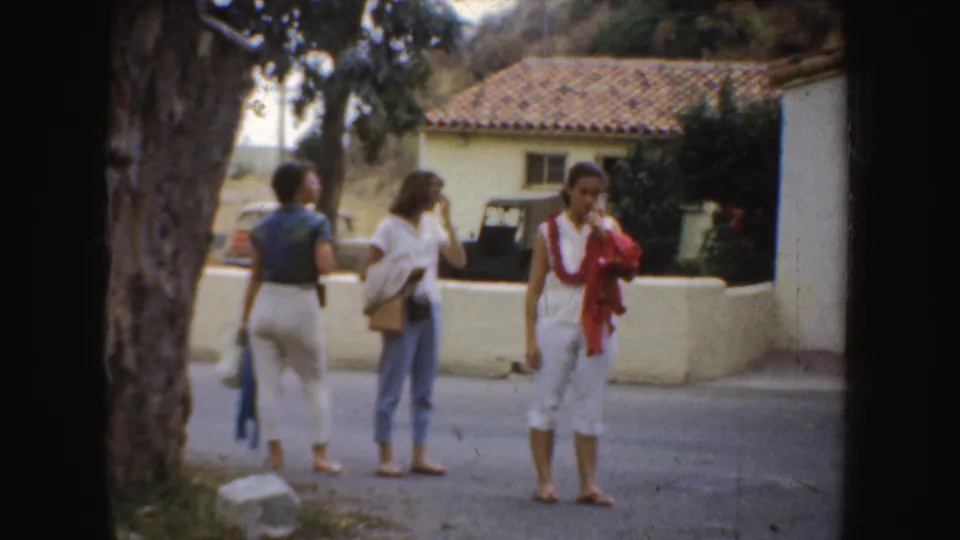 1952 California home-movie frame showing three women talking casually near a tree and tiled-roof exterior.