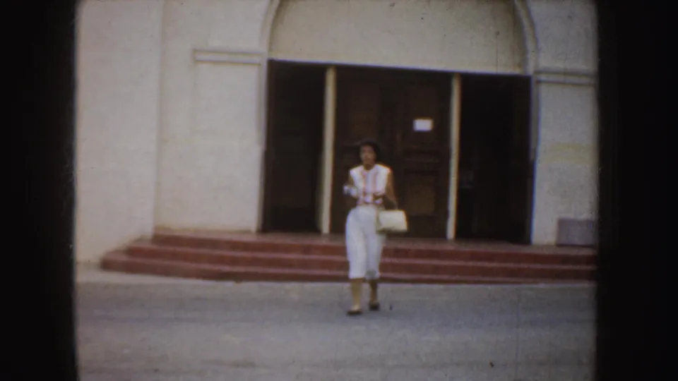 An adult woman in white clothing walks from an arched entrance with steps, likely part of the same California exterior scene.