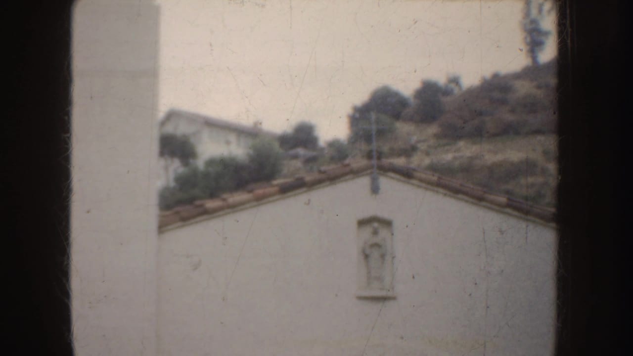 1952 California 8mm frame showing a white stucco mission-style wall, a small statue niche, red tile roofline, and hillside vegetation.