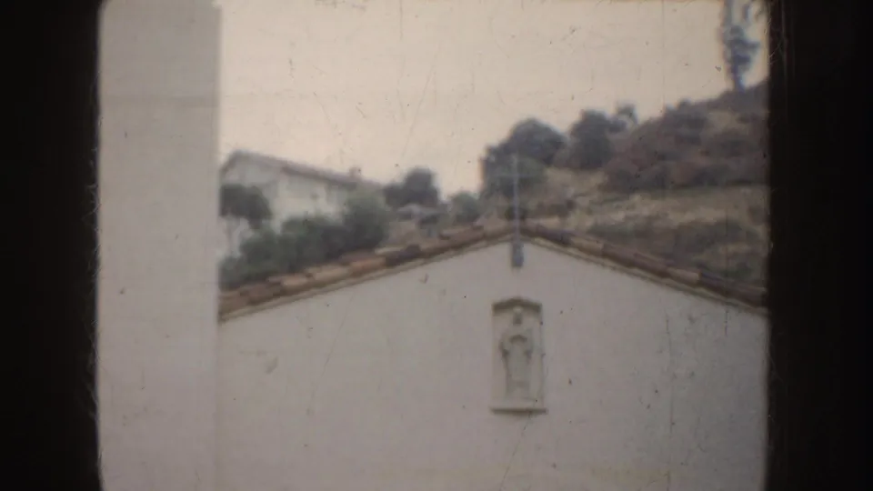 1952 California 8mm frame showing a white stucco mission-style wall, a small statue niche, red tile roofline, and hillside vegetation.