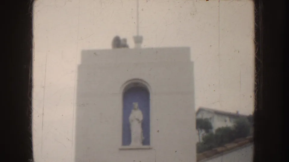 1952 California 8mm frame with a white stucco tower facade, arched blue recess, religious statue, rooftop details, and adjacent roofline.