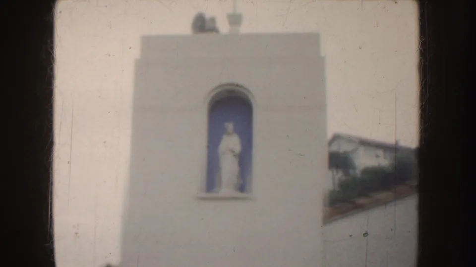 Soft 1952 California 8mm view of the mission-style tower, arched statue niche, pale stucco wall, and red tile roof beside it.