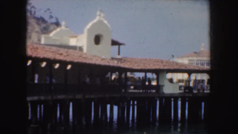 1952 California 8mm frame showing a covered pier, white mission-style roof forms with crosses, water, pilings, and small visitors in the distance.