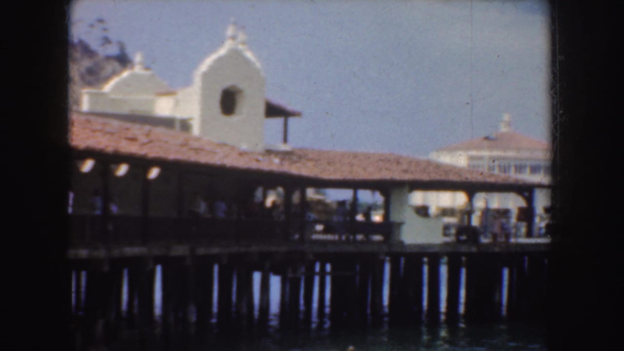 1952 California harbor view showing a covered pier, white waterfront building, dock pilings, and people in shadow.