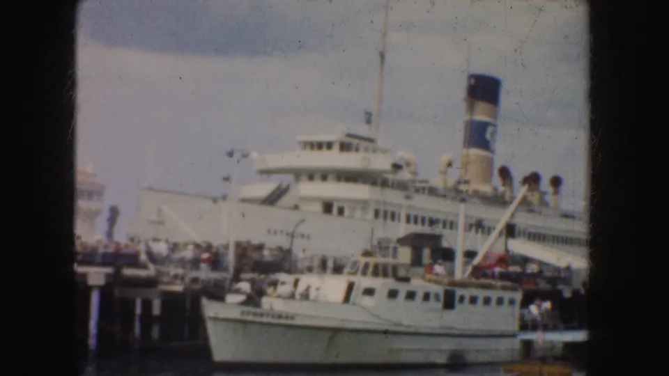 A large white passenger ship with a prominent smokestack sits at a California dock with a smaller boat in front.