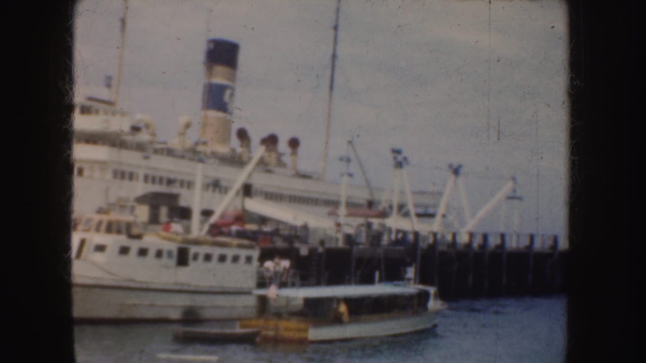 1952 California 8mm frame showing a large white passenger ship with a blue-and-white smokestack docked beside small harbor boats.