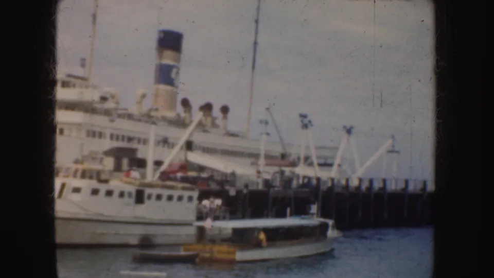 1952 California 8mm frame showing a large white passenger ship with a blue-and-white smokestack docked beside small harbor boats.