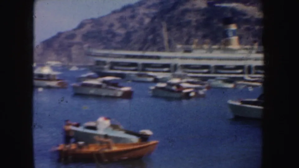 Soft 8mm view of a passenger ship near a rocky California harbor shoreline, with small motorboats clustered in the foreground.