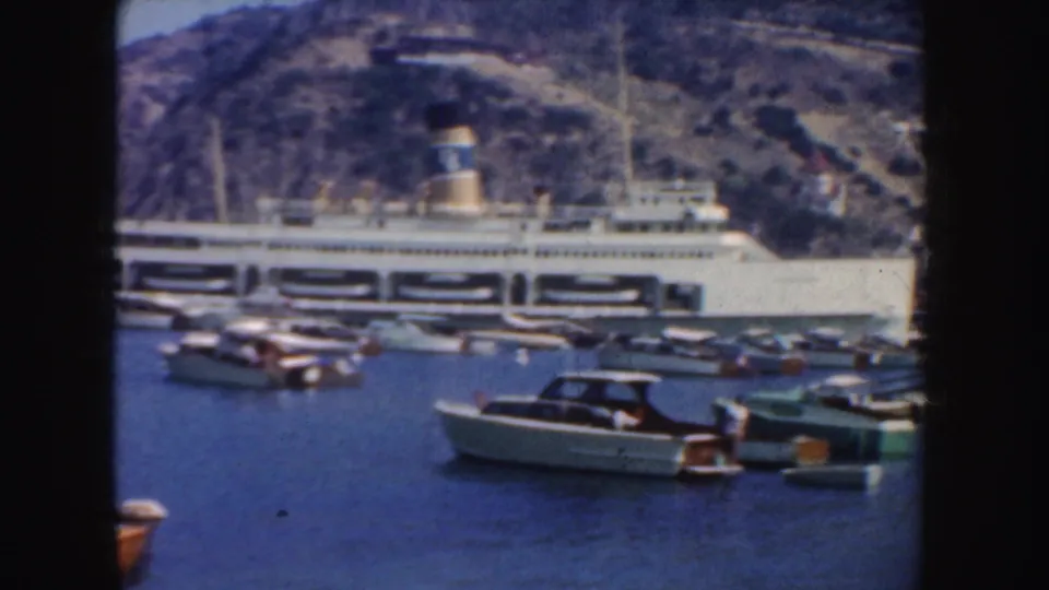 1952 California harbor frame with small motorboats in the foreground and a white passenger ship moored below a rocky hillside.
