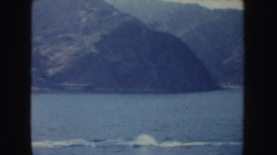 A white wake trails across blue water with rugged California shoreline in the distance, filmed on soft 8mm home-movie stock.