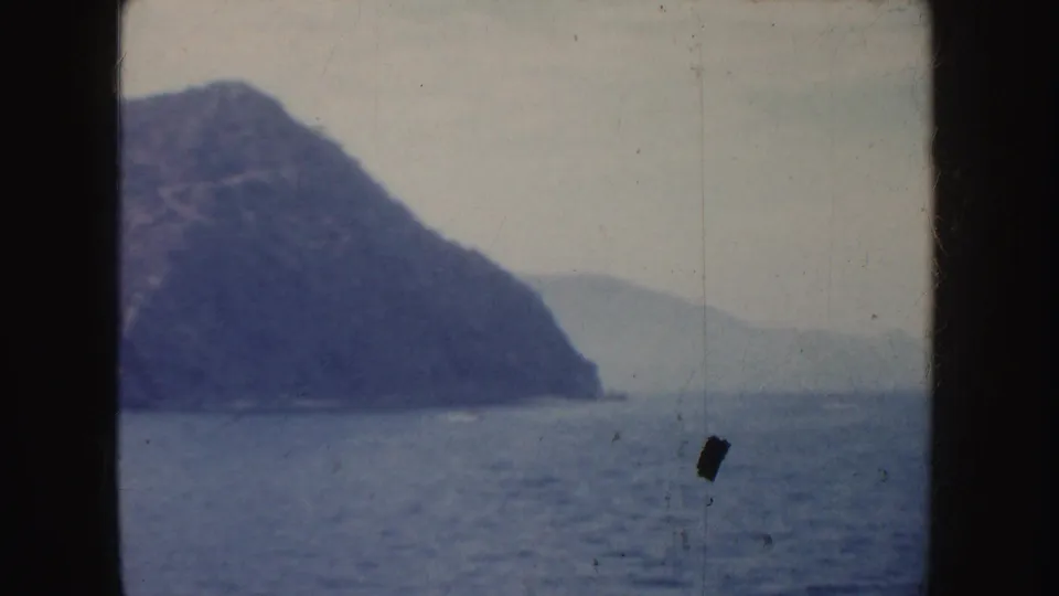 Open water and rugged California shoreline with a small indistinct towed figure or object near the right edge, captured in 1952 home-movie footage.